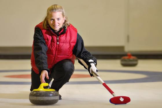 A woman kneels on the ice with a curling stone and broom
