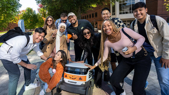 Students group up for a selfie with a starship robot that has been decorated with a "pumkinbot" halloween skin.