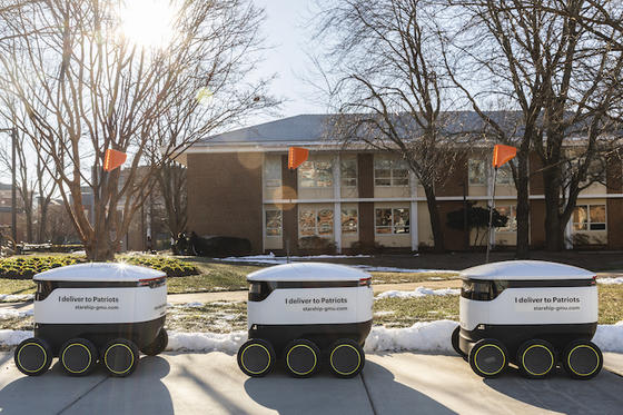 Three Starship robots sit queued on the sidewalk outside some academic buildings on the Fairfax Campus.