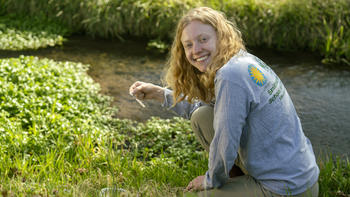 Student Maggie Walker sits next to a jet of water and smiles at the camera.  Wearing an SMSC shirt, she holds a QuanTab strip to measure chloride levels in the stream.