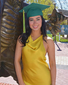 A young woman in a gold dress and a green graduation cap stands next to a statue.