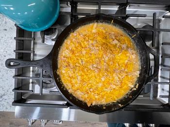 A cast iron skillet full of white chicken chili, with cheddar cheese sprinkled on top, sitting on the burner of a gas range.