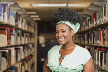 Headshot of Amini Bonane. She is standing in between aisles of books at the library.