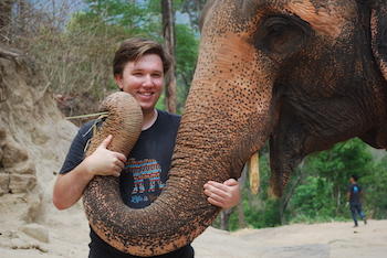 Chase LaDue smiling. He is standing near an elephant with both hands on its trunk. The end of the elephant's trunk is by Chase's face.