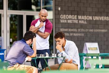 Two men are sitting together at a table on the plaza at Mason Square playing chess. A third man is standing next to the table looking at the game board. In the background, the front of the building says George Mason University