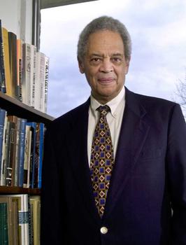 Roger Wilkins stands for a portrait in a sunlit office with full bookshelves. He is wearing a suit and tie, and smiles gently.