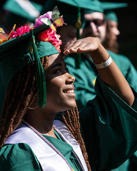 A graduate shields her face from the sun with her hand as she stands in line with other graduates. Her graduation cap is decorated with silk butterflies and flowers.