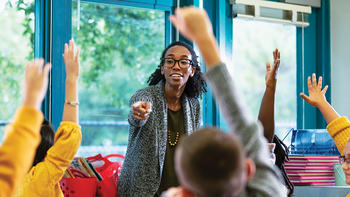 Stock image of a teacher pointing at a student out of a group. Many of the students have their hands raised to answer a question.