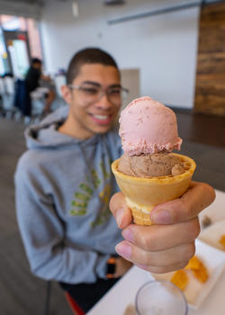 A student holds up a strawberry and chocolate gelato in a cone for the camera.