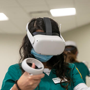 A student in nursing scrubs is wearing a VR headset and using two VR controllers to interact with the program inside the headset.