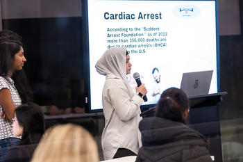 Dallia Laban wears a beige hijab and speaks into a microphone. She stands in front of a slide with statistics on cardiac arrest rates.