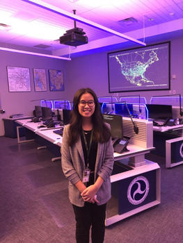 A young woman with long hair wearing glasses stands in a room full of computers.