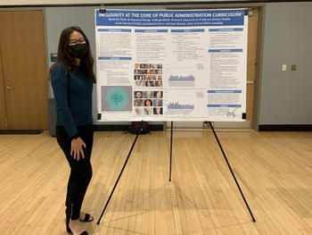 A young woman wearing a mask stands next to a research poster.